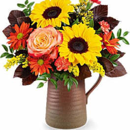 Bright arrangement of sunflowers, roses, and orange daisies in a rustic brown pitcher