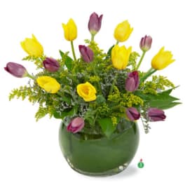 Arrangement of yellow and purple tulips with yellow filler flowers in a round clear glass vase