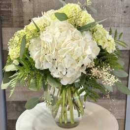 White and pale green hydrangea bouquet with lush foliage in a clear glass vase on a round white stand