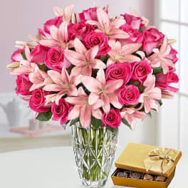 Pink roses and lilies in a glass vase beside a box of chocolates