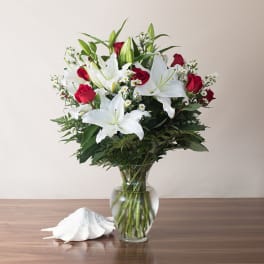 Tall arrangement of white lilies, red roses, and small white blooms in a clear glass vase on a table