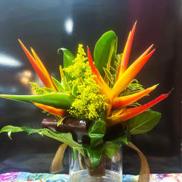Compact tropical arrangement with orange and yellow flowers and bold green leaves in a clear glass vase.