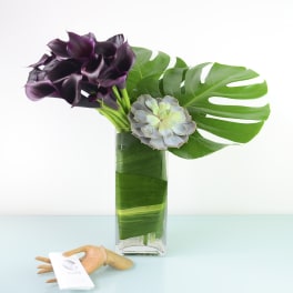 Purple calla lilies and a succulent in a glass vase with large tropical leaves