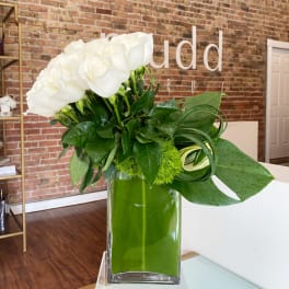 White roses arranged in a tall glass vase with green foliage.