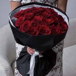 Large bouquet of deep red roses wrapped in black paper with a white ribbon, held by a person.