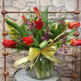 Mixed bouquet of red tulips and small purple flowers in a clear glass vase with yellow ribbon.