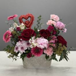 Pink and red mixed flower arrangement in a decorative white container with a glitter heart pick
