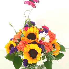 Mixed bouquet with sunflowers, roses, and daisies in a glass vase topped with a butterfly