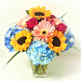 Bouquet of sunflowers, pink gerberas, roses, and blue hydrangeas in a clear vase