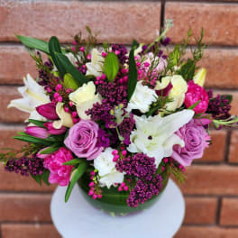 Round arrangement of pink, purple, and white blooms in a leaf-lined glass vase.
