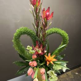 Tall floral arrangement with pink and orange blooms in a gold vase, framed by green heart shapes.