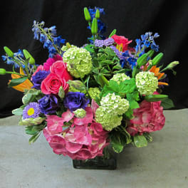 Colorful arrangement of pink hydrangeas, roses, green blooms and mixed purple and blue flowers in a glass cube vase