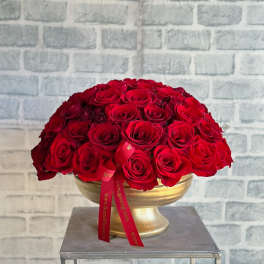 Round arrangement of red roses in a gold pedestal bowl with a red ribbon