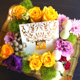 Floral gift arrangement with yellow roses, purple blooms, and decorative eggs in a tray