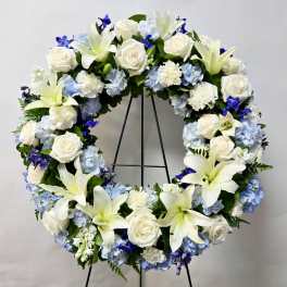 Large floral wreath with white lilies, roses, and blue hydrangeas on a stand