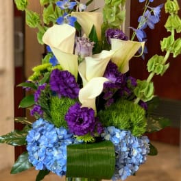 Tall bouquet of white calla lilies, blue flowers, and purple blooms in a glass vase