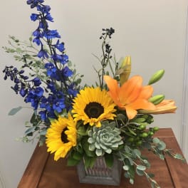 Bright mixed flower arrangement in a wooden box with sunflowers, lilies, and blue blooms