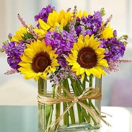Sunflowers and purple flowers in a clear glass vase with a raffia bow