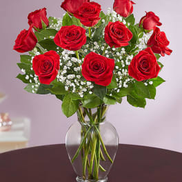 Dozen red roses with white filler flowers arranged in a clear glass vase on a dark table
