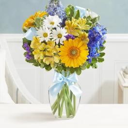 Mixed bouquet of daisies, gerberas, and blue flowers in a glass vase with a ribbon