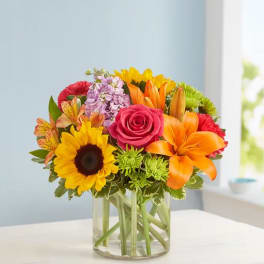 Bright mixed bouquet in a clear glass vase with sunflower, rose, lily, and orange blooms