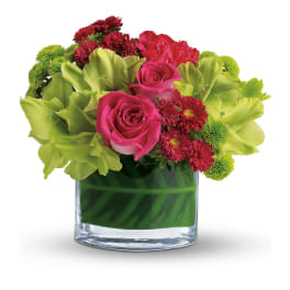 Low arrangement of hot pink roses, red mums, and green blooms in a clear glass cylinder vase