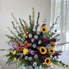 Large mixed floral arrangement with sunflowers, roses, and purple blooms in a pedestal vase