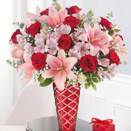 Bouquet of pink lilies and red roses in a red vase with a gift box nearby