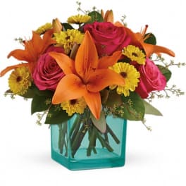 Bright orange lilies, pink roses, and yellow blooms in a turquoise glass cube vase
