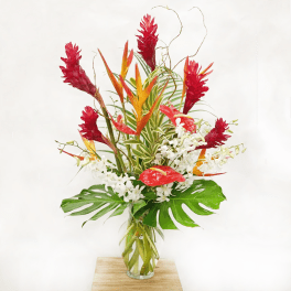 Large tropical arrangement with red and orange blooms, white orchids, and bold green monstera leaves in a glass vase