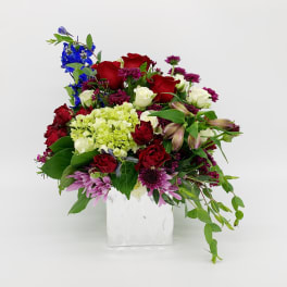Mixed red, white, green, and purple flower arrangement in a white square vase with trailing foliage