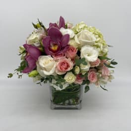 Compact arrangement of pink and white roses with purple orchids in a clear glass cube vase