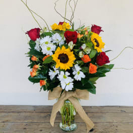 Tall mixed bouquet with sunflowers, red roses, white daisies, and orange blooms in a glass vase with burlap bow