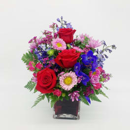 Vibrant mixed bouquet with red roses, pink daisies, and blue flowers in a red glass cube vase