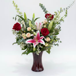 Tall arrangement of red roses, pink lilies, and peach spray roses in a dark red glass vase