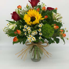 Mixed bouquet with a sunflower, red and orange roses, daisies, and white blooms in a glass vase with raffia bow.