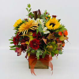 Square wooden vase arrangement with yellow sunflowers, red mums, white blooms, and orange accents.