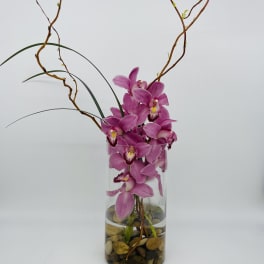 Tall pink orchid arrangement in a clear glass cylinder vase with river stones at the base