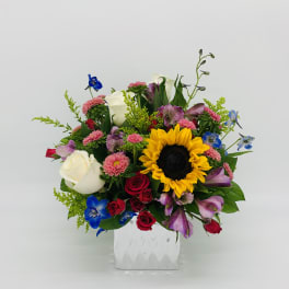Colorful mixed bouquet with sunflower, roses, mums, and blue blooms in a white glass cube vase