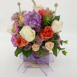 Mixed bouquet of pink, orange, and white roses with purple blooms in a wood box with lavender bow.