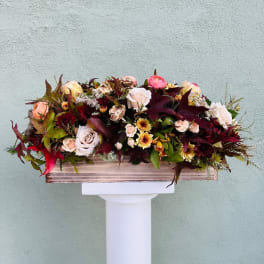 Low rectangular arrangement of blush roses and autumn-toned flowers in a wooden box on a white pedestal.