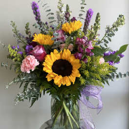 Tall mixed arrangement with sunflowers, pink roses, and purple flowers in a clear glass vase