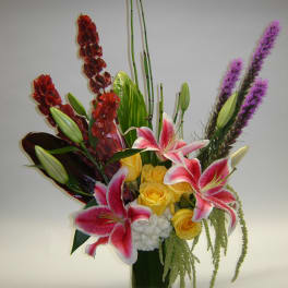 Mixed bouquet with pink lilies, yellow roses, and purple spikes in a glass vase