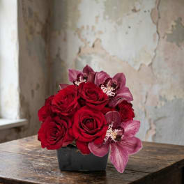 Low arrangement of red roses and mauve orchids in a black square vase on a rustic wooden table.