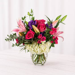 Bouquet of pink roses, lilies, and purple blooms in a clear glass vase