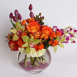 Mixed bouquet of orange roses, purple tulips, and orchids in a round glass vase