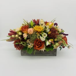 Low centerpiece of orange roses, yellow mums, and mixed flowers in a rustic wooden box.