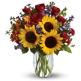Bouquet of sunflowers, red roses, and purple filler flowers in a clear glass vase