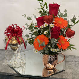 Red roses and orange gerbera daisies in a copper mug beside a gift jar of chocolates