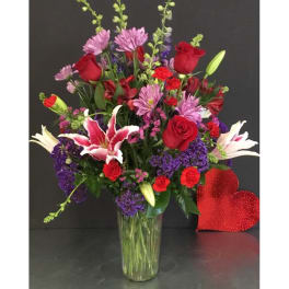 Tall mixed arrangement of red roses, pink lilies, and purple flowers in a clear glass vase.